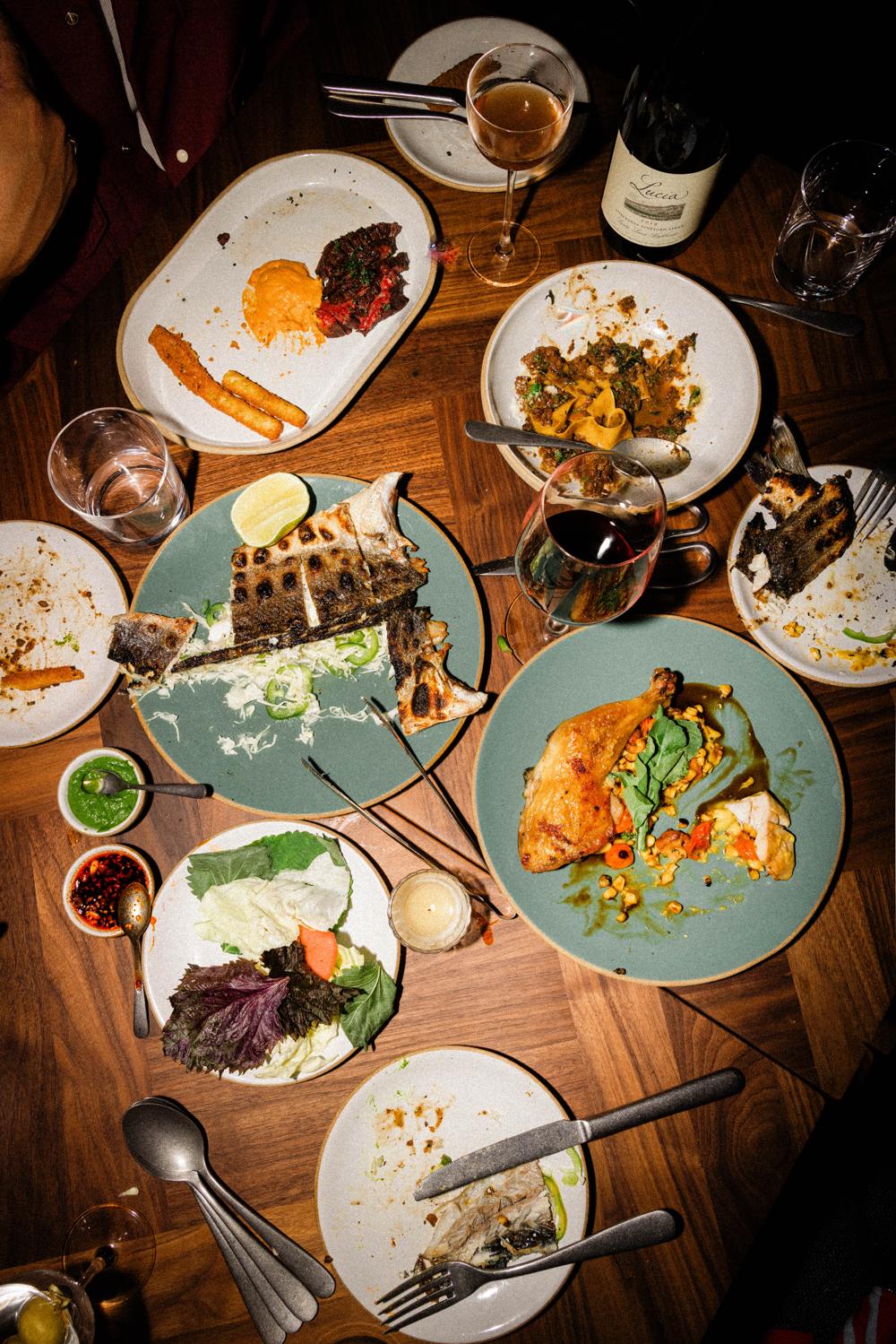 A family-style spread of branzino, half-chicken, steak, salads and wine across the table